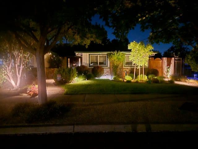 Illuminated suburban home at night with landscaped garden and trees along the sidewalk.