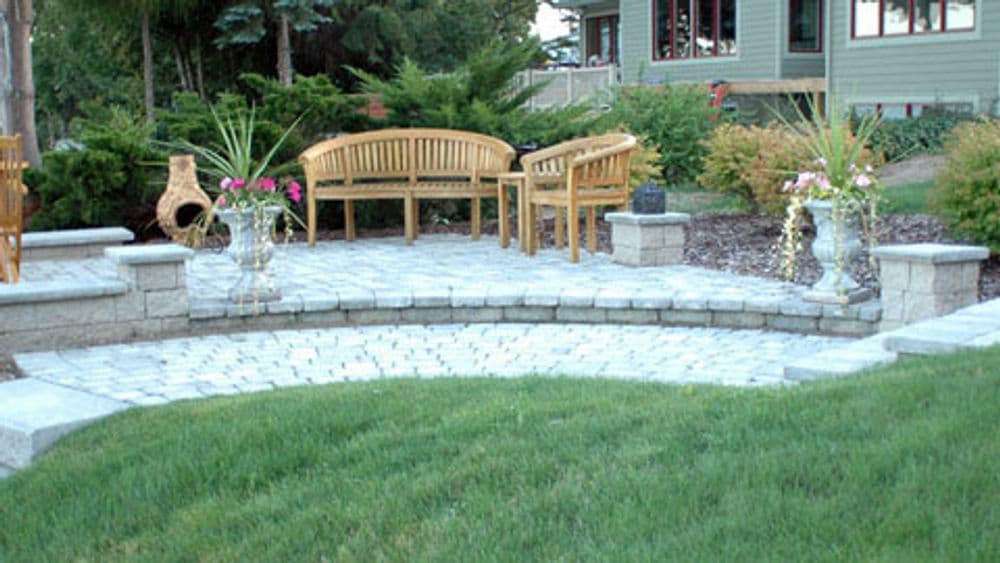 Patio area with stone pavers, wooden benches, and garden planters surrounded by greenery.