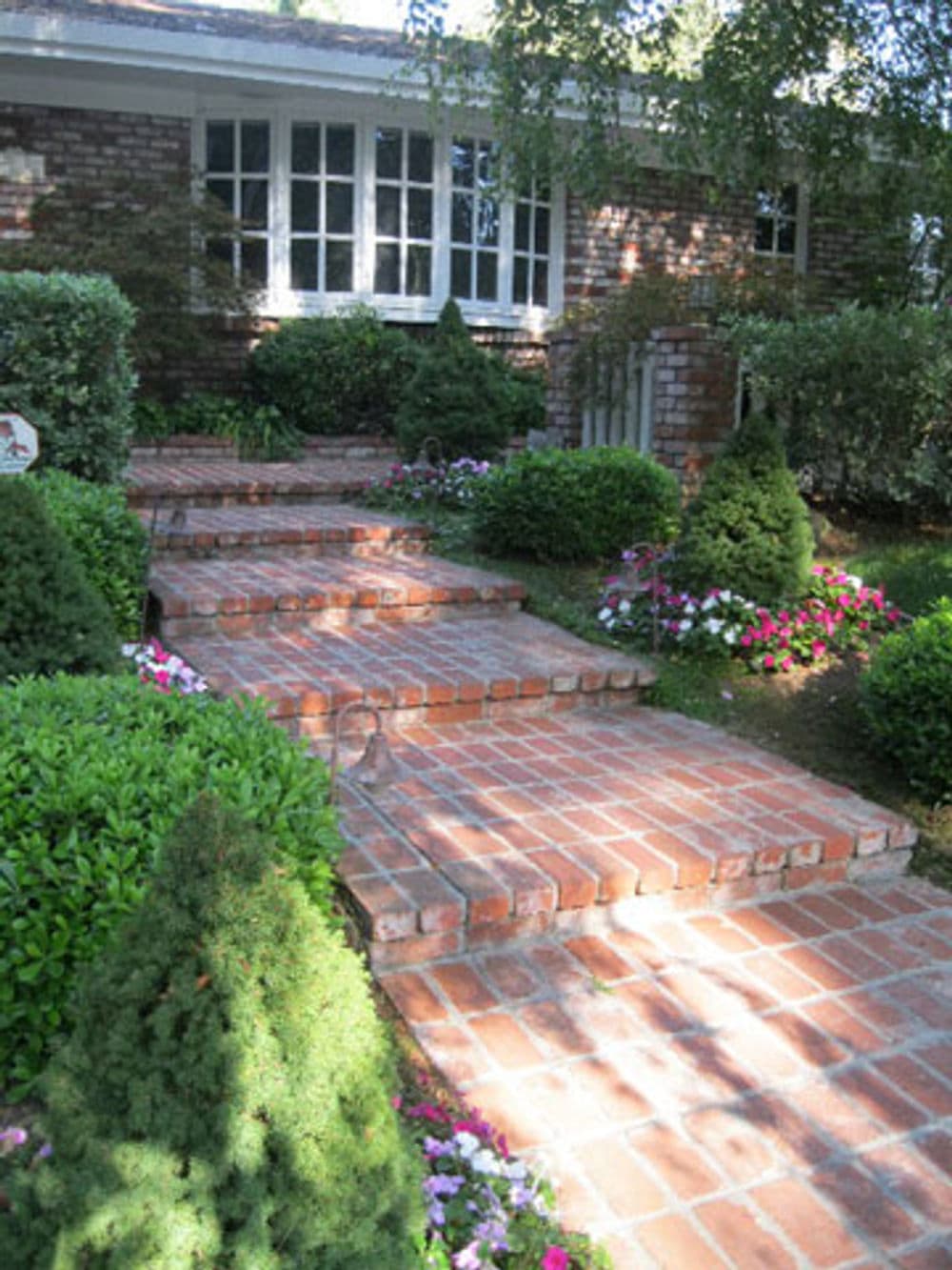 Brick pathway leading to a house, surrounded by green shrubs and colorful flowers.