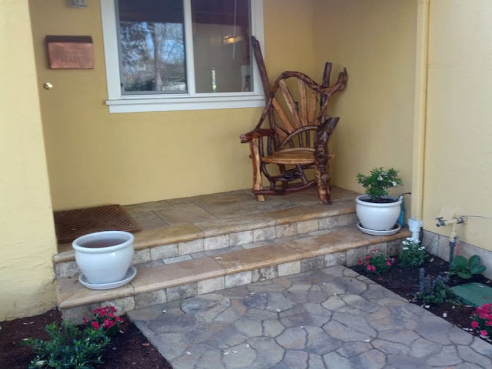 Rustic wooden chair on a stone porch with potted plants and yellow wall backdrop.