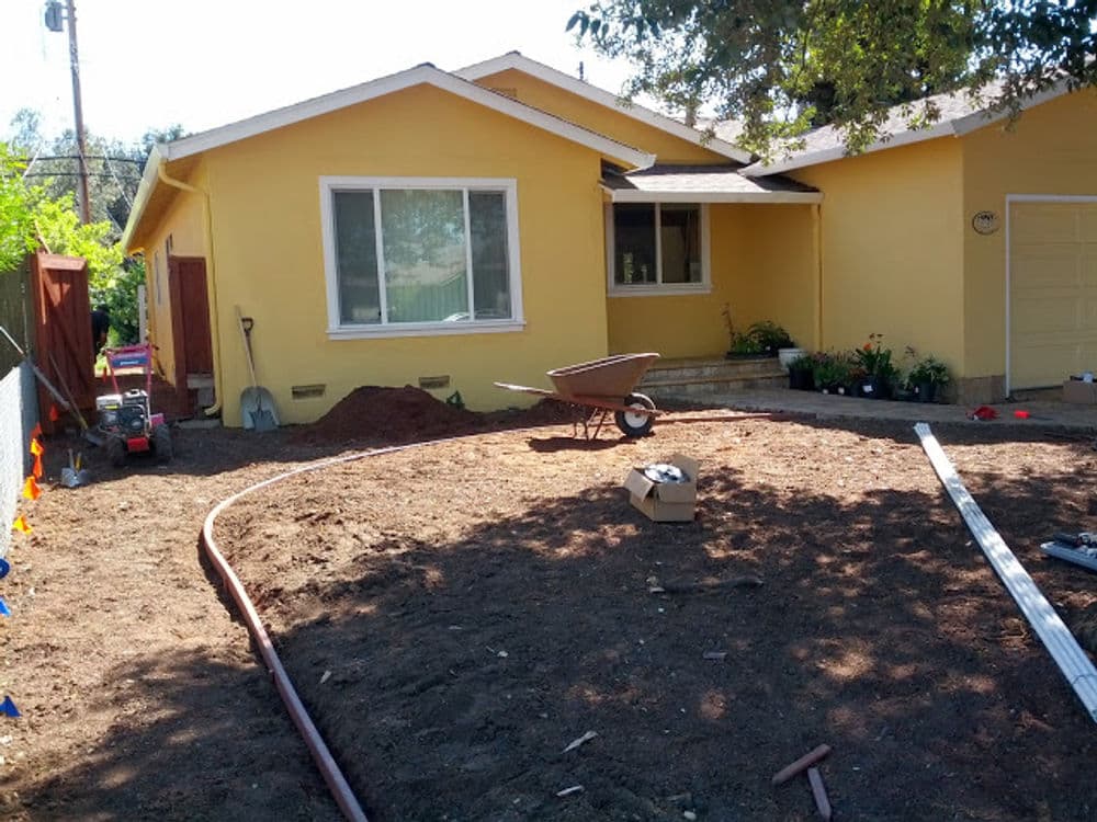 Residential yard renovation with yellow house, wheelbarrow, and garden preparation materials.
