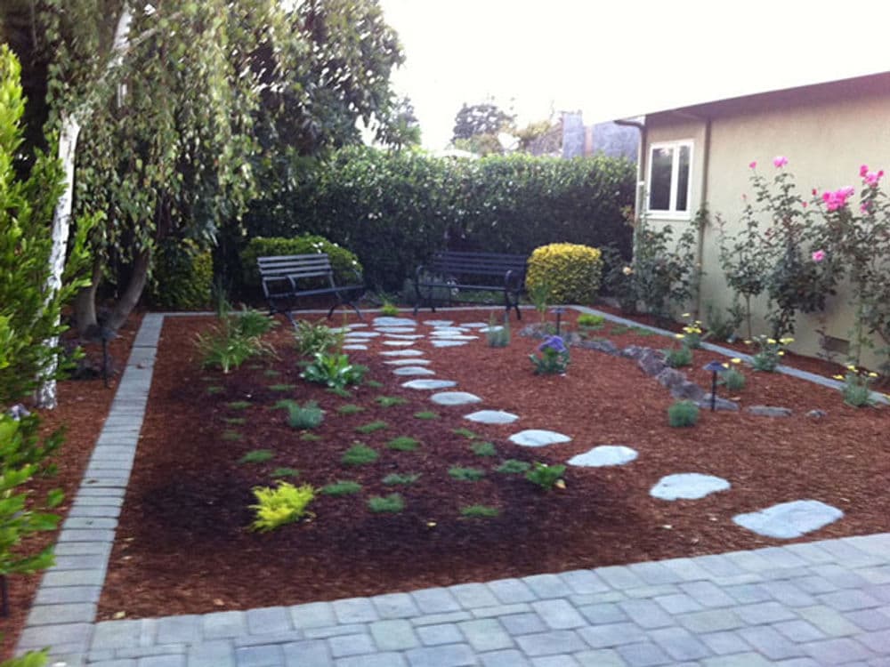 Lush garden with stone pathway, benches, and flowering plants in a serene backyard setting.