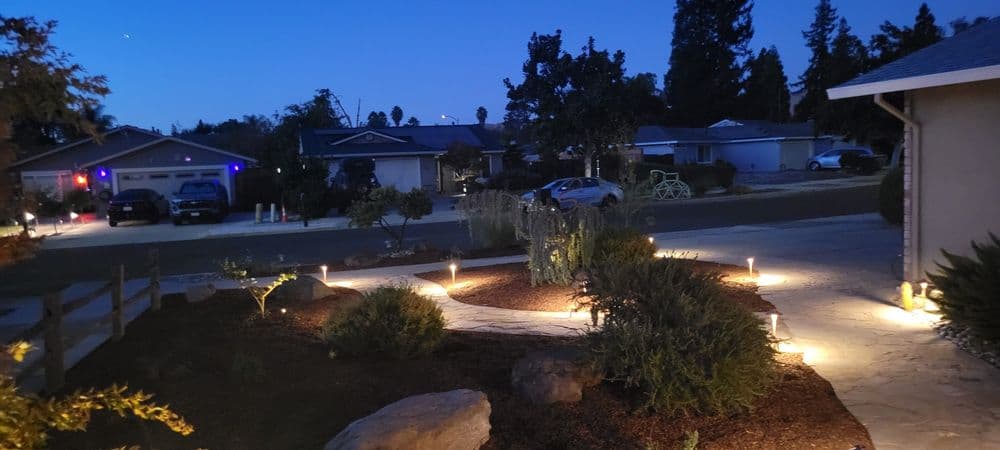 Illuminated garden pathway at dusk with landscaped plants and neighboring homes in the background.